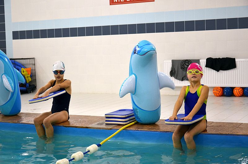Dwie dziewczynki w strojach kąpielowych i czepkach siedzą na brzegu basenu, trzymając deski do pływania; obok nich dmuchany delfin i piłki do ćwiczeń.