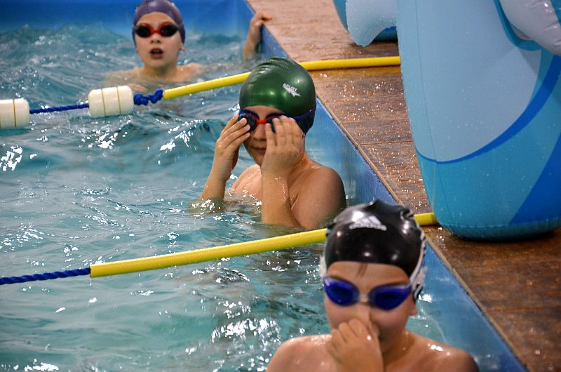 Trójka dzieci w czepkach i okularach pływackich w basenie, jedno poprawia okulary, widoczna lina oddzielająca tory i dmuchana zabawka delfin w tle.