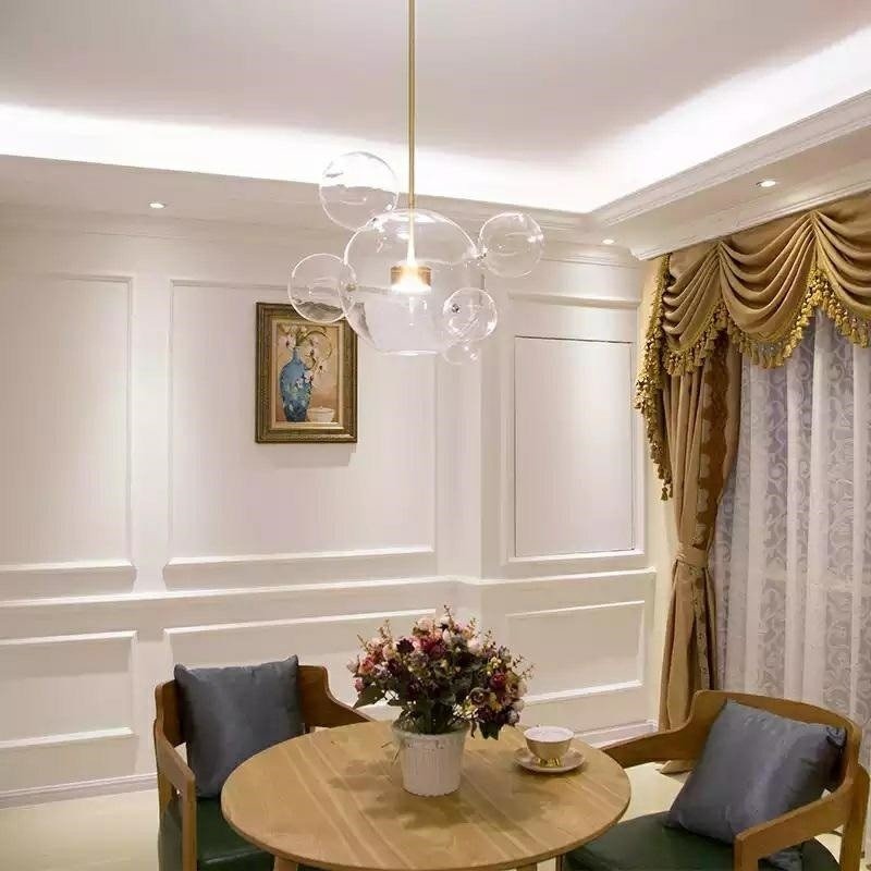 Nowoczesna lampa sufitowa z przezroczystych kul w eleganckim salonie z dekoracyjnymi sztukateriami, kwiatami na okrągłym stole i zasłonami w kolorze złota.