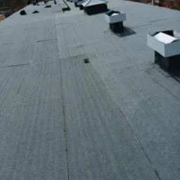 Widok z góry na płaski dach pokryty papą termozgrzewalną w Kolnie, z kominami wentylacyjnymi i ekipą dekarzy w trakcie pracy, widoczny budynek w tle.