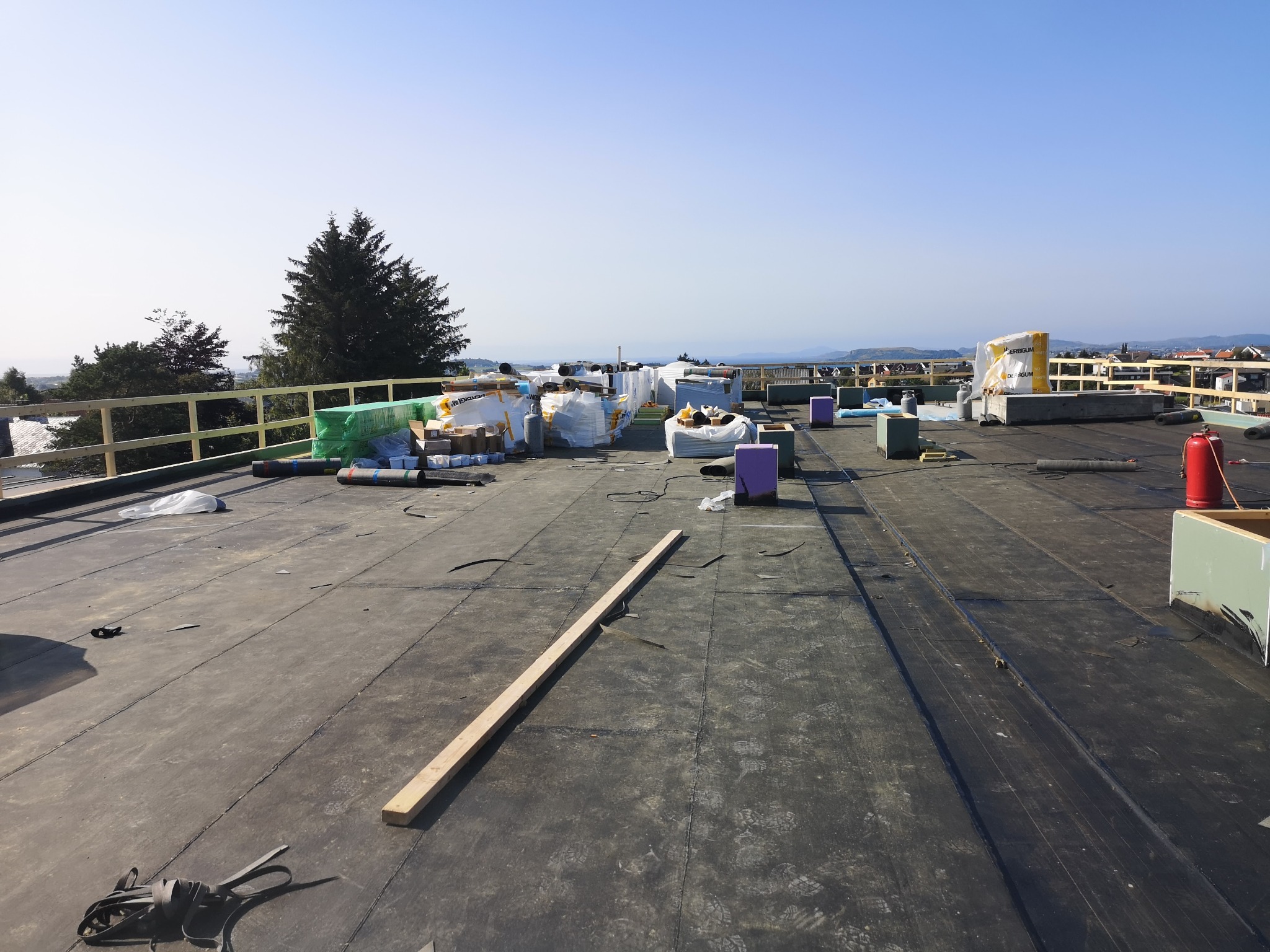 Widok na płaski dach pokryty papą, z widocznymi materiałami budowlanymi, narzędziami, oraz elementami zabezpieczeń na krawędzi dachu, z panoramą miasta i wzgórz w tle.