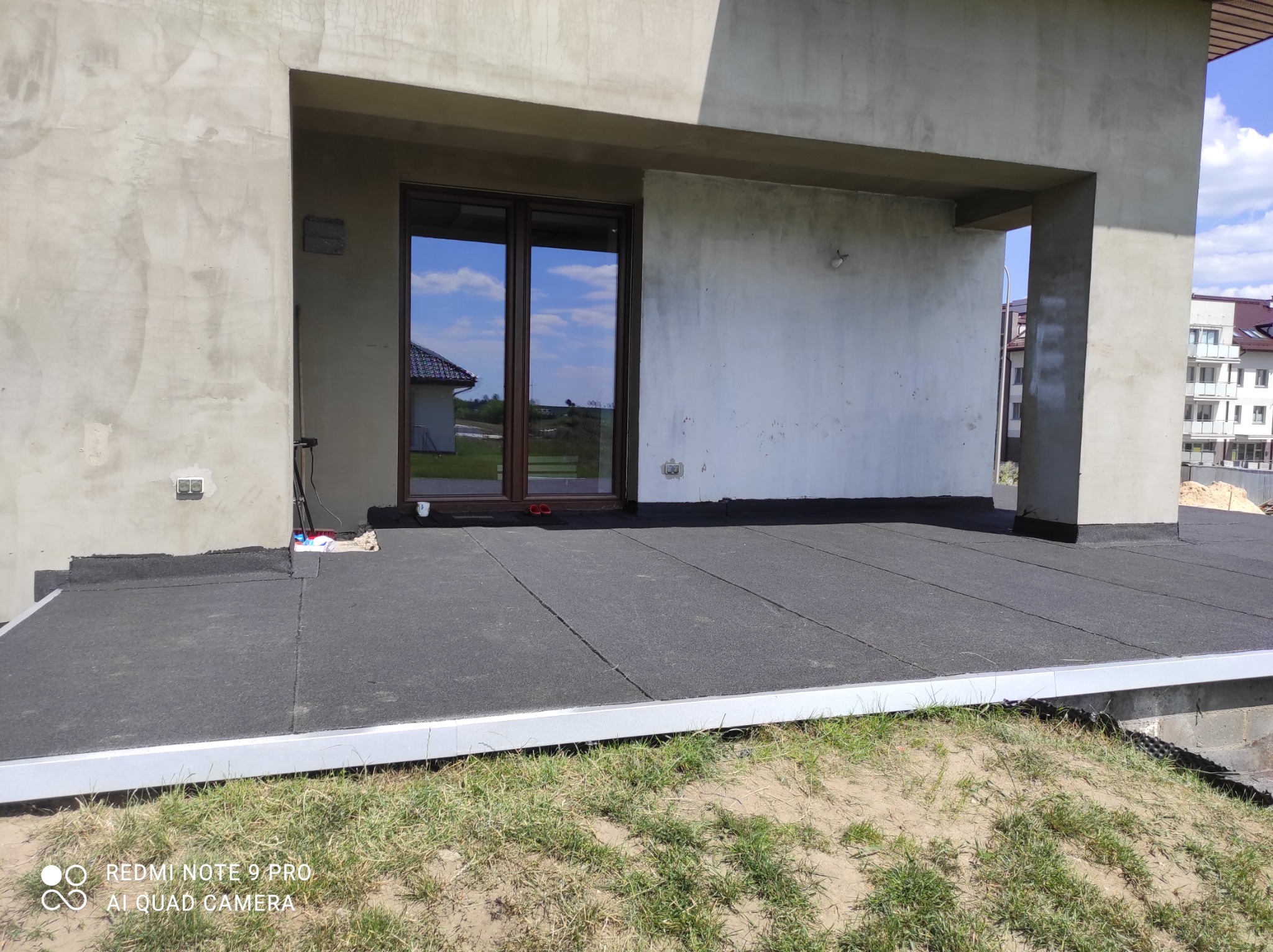 Wykończony płaski dach budynku pokryty czarną papą termozgrzewalną, z widocznymi detalami obróbek blacharskich przy krawędzi dachu i oknach balkonowych, w tle budynki mieszkalne i błękitne niebo...