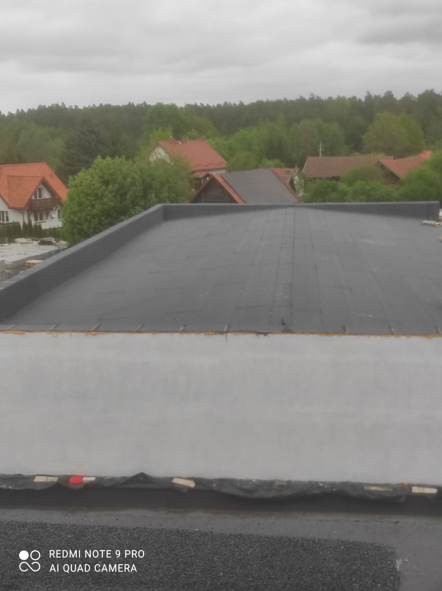 Płaski dach pokryty czarną papą termozgrzewalną, widoczne łączenia papy, w tle zabudowa jednorodzinna i las pod pochmurnym niebem.