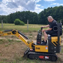 Operator obsługuje żółtą mini koparkę NEX N10 na trawiastym terenie z widocznym lasem w tle pod pochmurnym niebem.
