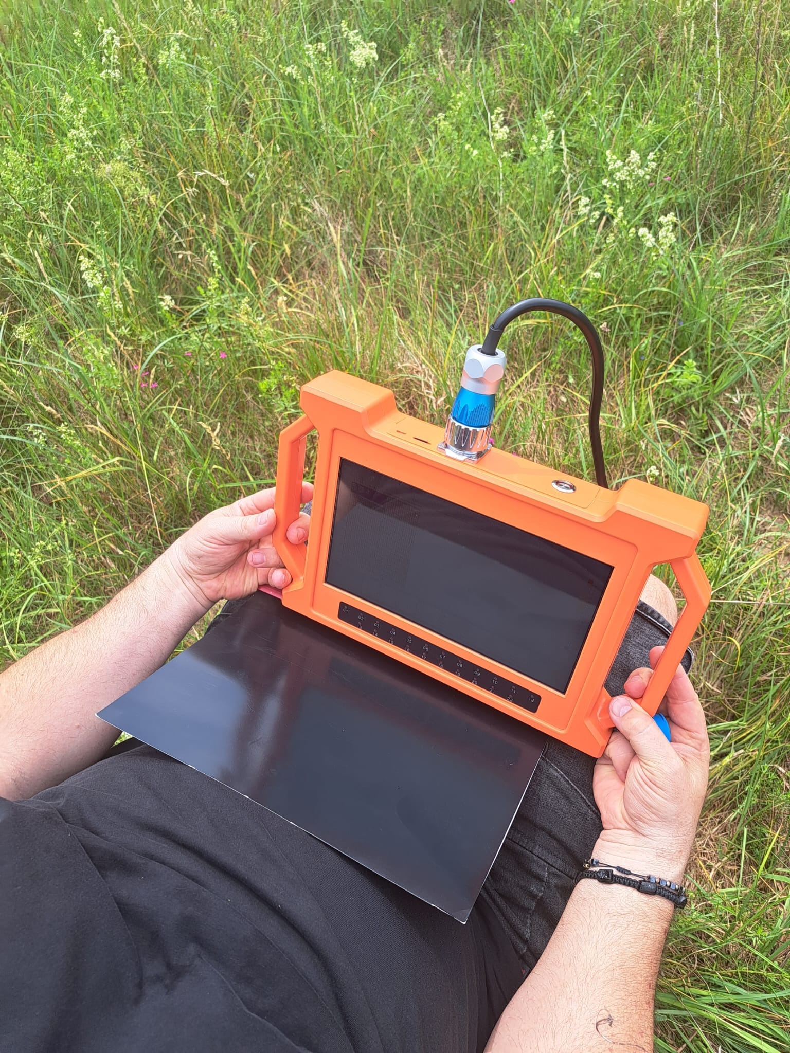 Osoba trzyma pomarańczowy monitor z podłączonym kablem, używany do inspekcji podziemnych instalacji; w tle trawa i polne kwiaty.