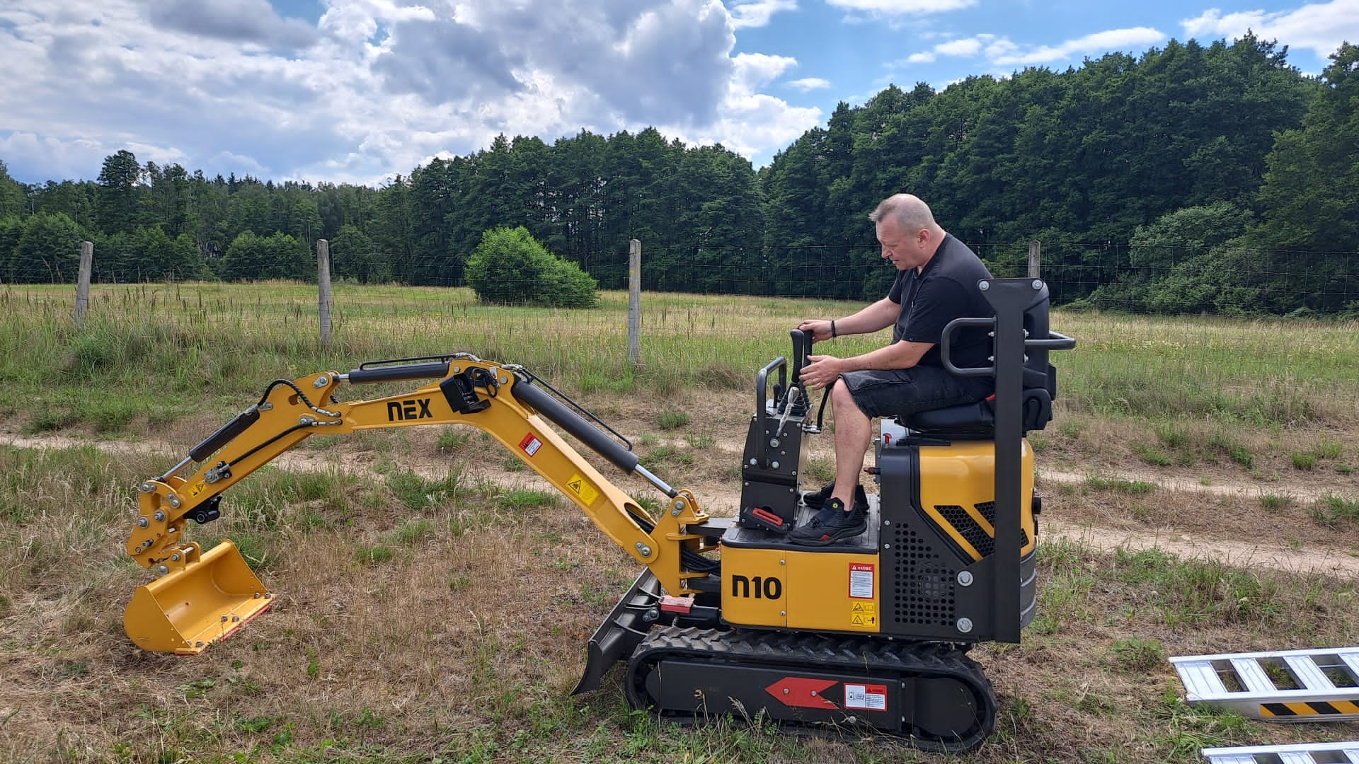 Operator obsługuje żółtą mini koparkę NEX N10 na trawiastym terenie z widocznym lasem w tle pod pochmurnym niebem.