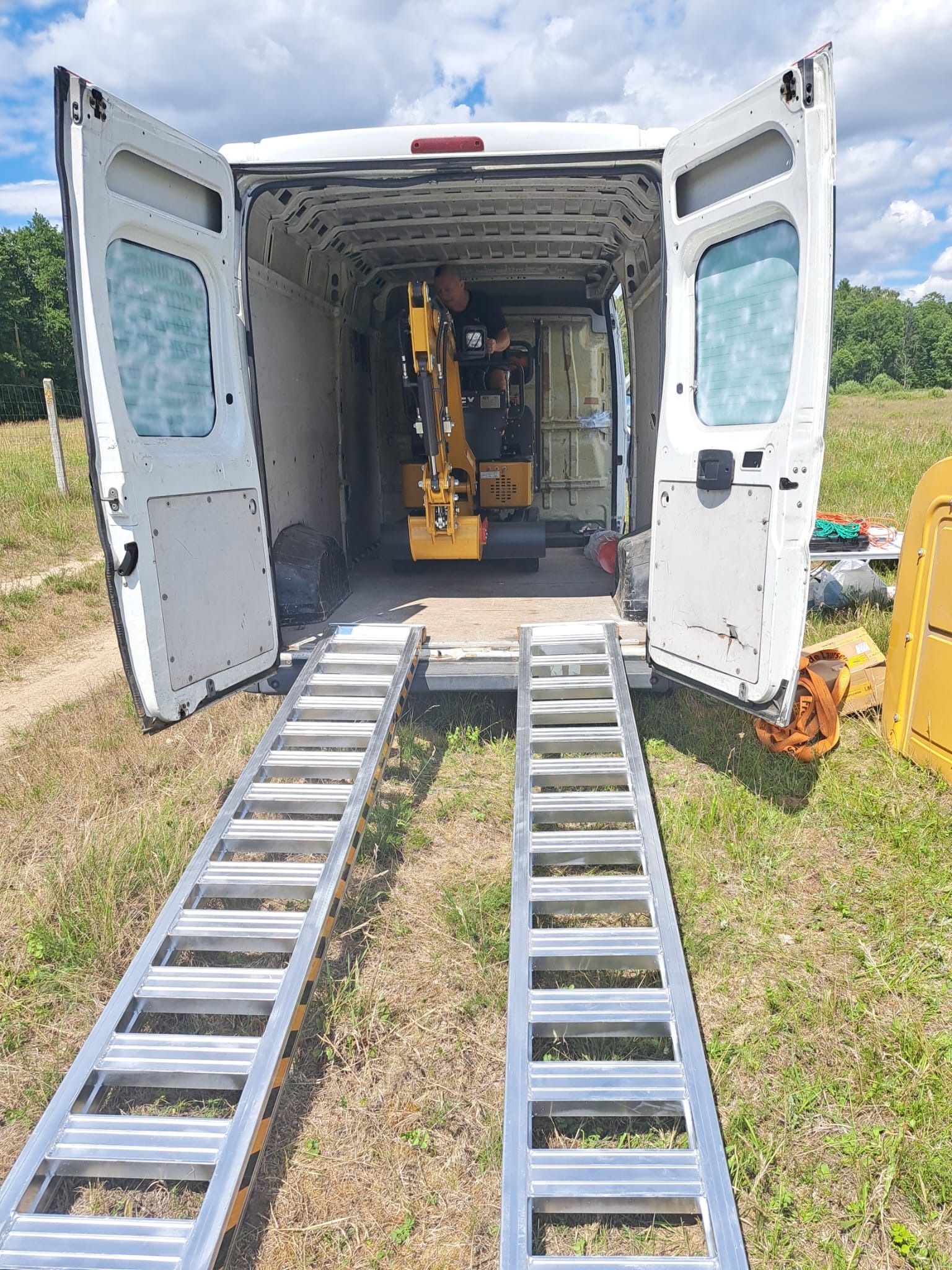 Żółta mini koparka na rampie wewnątrz białego samochodu dostawczego z otwartymi drzwiami, widok z zewnątrz na łąkę i las.