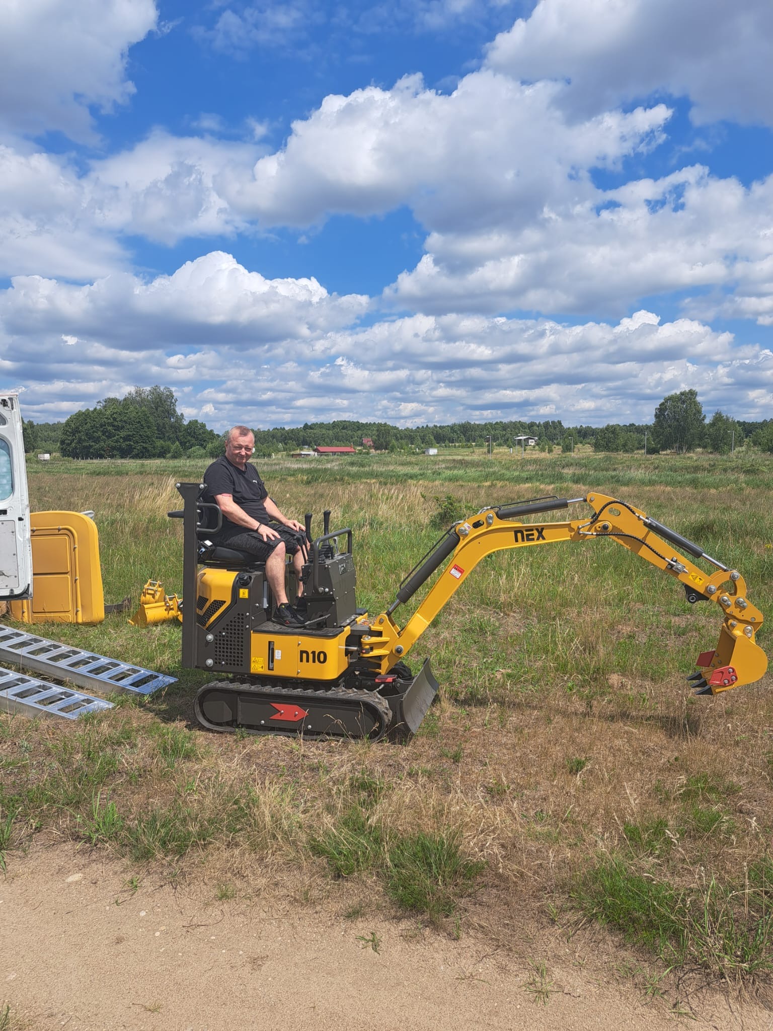 Operator siedzi na żółtej mini koparce N10 na tle łąki i błękitnego nieba z chmurami. Rampa najazdowa i przyczepa obok.