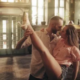 Para tańczy w jasnym, klimatycznym wnętrzu z dużymi oknami, kobieta w różowej bluzce i jeansowych szortach unosi nogę w beżowym bucie na obcasie, mężczyzna w jasnej koszuli ją podtrzymuje.