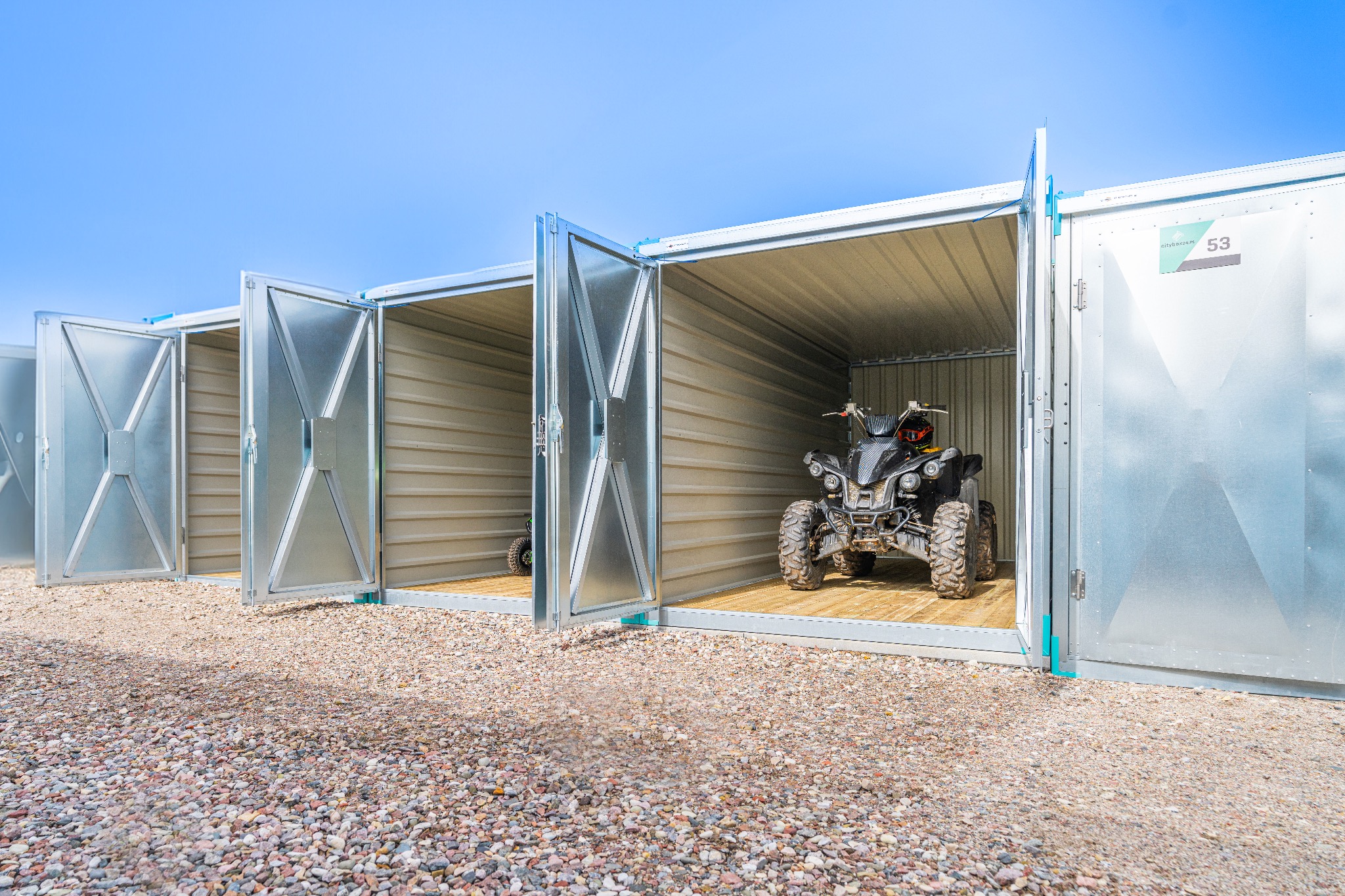 Rząd otwartych, metalowych boksów do przechowywania, z quadem zaparkowanym w jednym z nich. Podłoże wysypane drobnymi kamieniami.