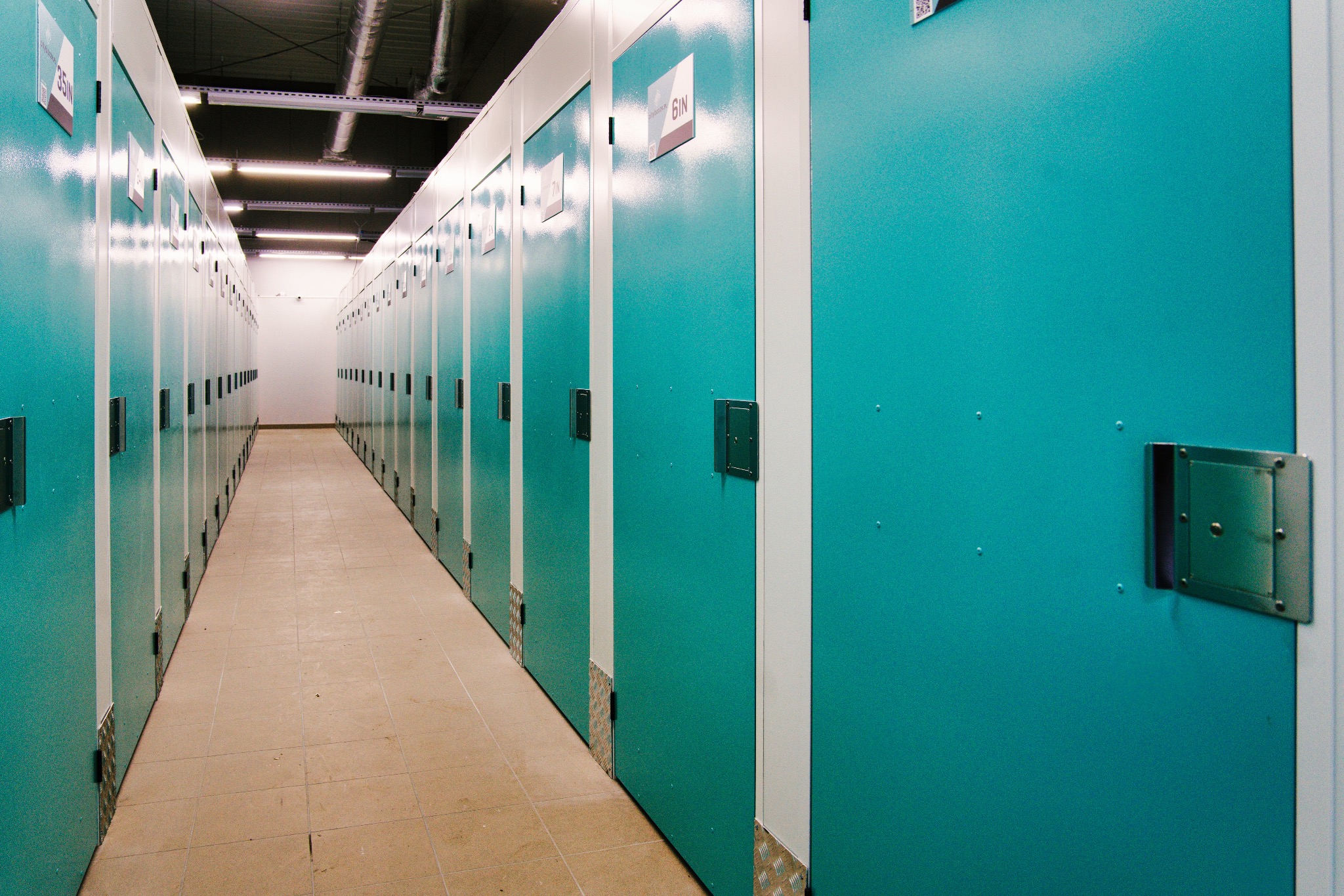 Korytarz z rzędami turkusowych boksów do przechowywania w Koszalinie, widok z perspektywy podłogi na wprost, oświetlenie sufitowe.