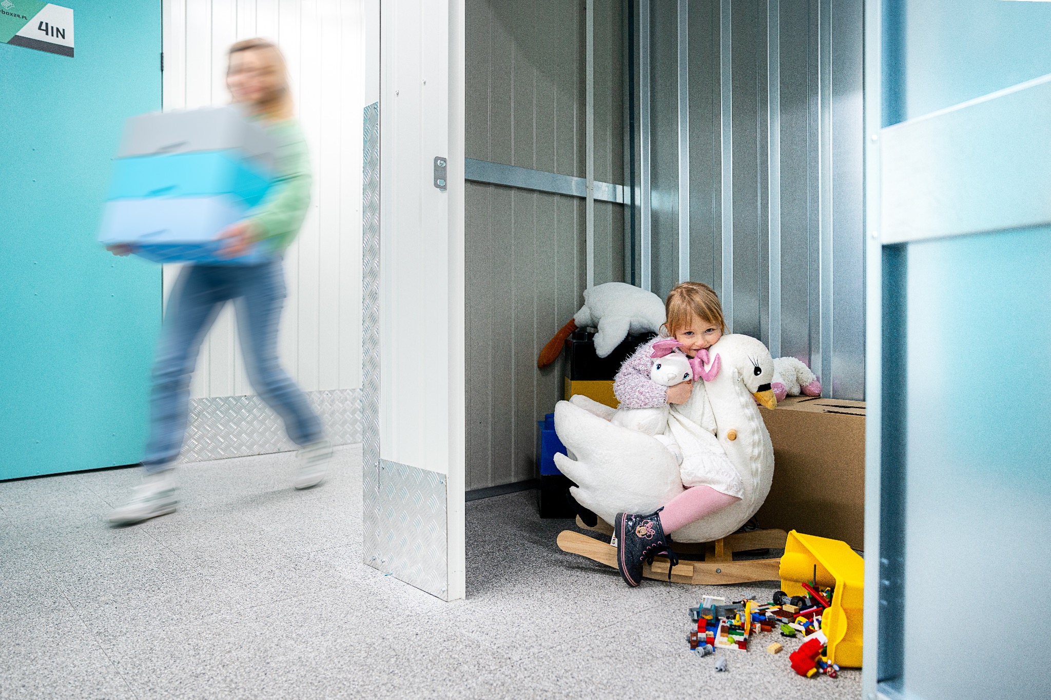 Kobieta w ruchu przenosi stos niebieskich pudeł w korytarzu z boksami; w otwartym boksie dziewczynka siedzi na bujanym łabędziu otoczona zabawkami.