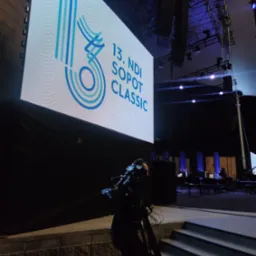 Operator kamery z profesjonalnym sprzętem wchodzi po schodach, w tle scena z ekranem LED wyświetlającym logo '13. NDI Sopot Classic' oraz elementy nagłośnienia i oświetlenia.