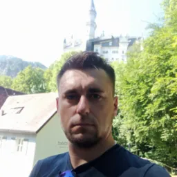 Mężczyzna robiący selfie na tle zamku Neuschwanstein, w tle zieleń drzew i błękitne niebo.