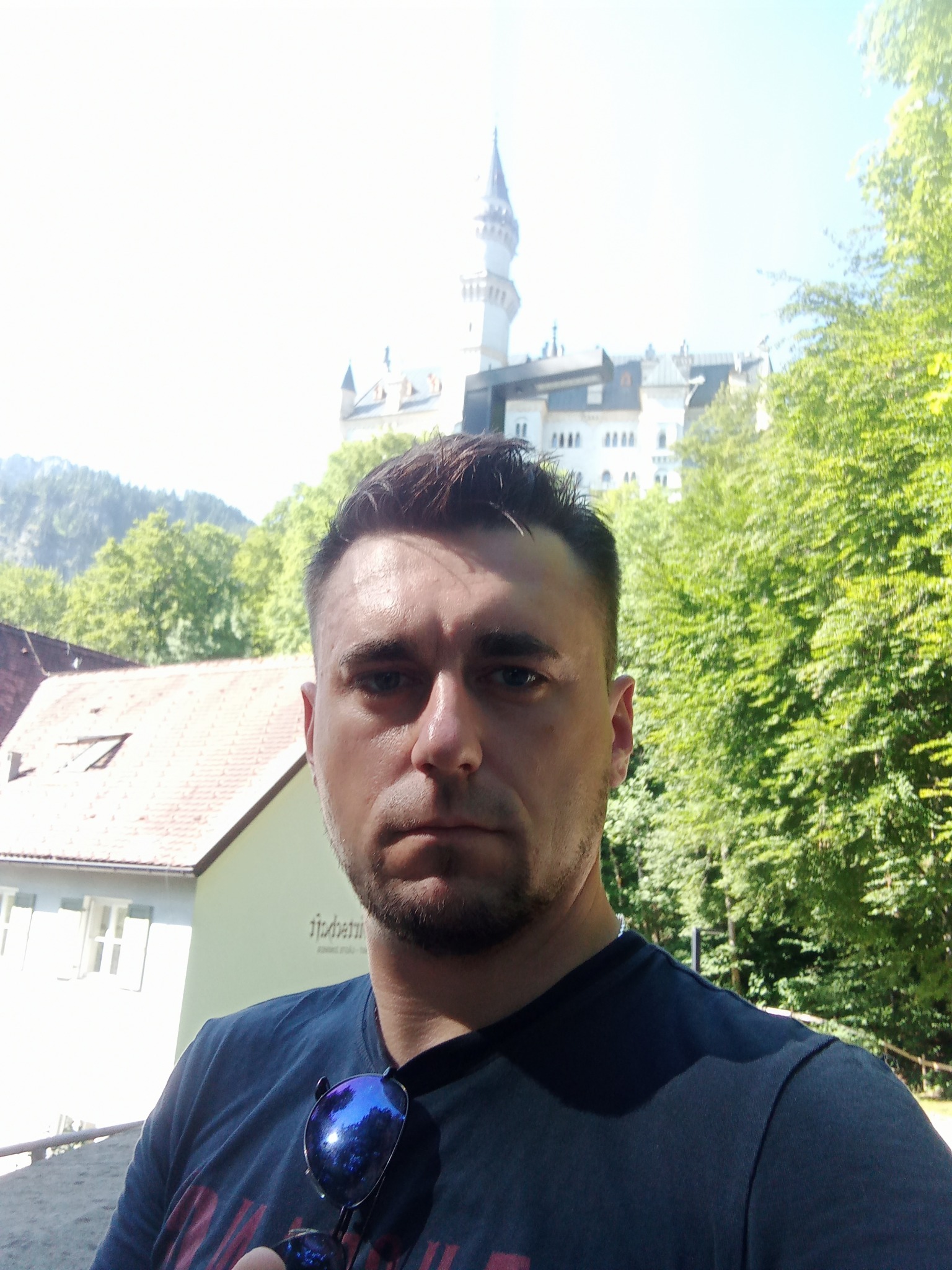 Mężczyzna robiący selfie na tle zamku Neuschwanstein, w tle zieleń drzew i błękitne niebo.