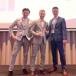 Trzech elegancko ubranych mężczyzn na scenie, jeden z nich trzyma statuetkę, na podłodze konfetti, w tle logo firmy Develocus.