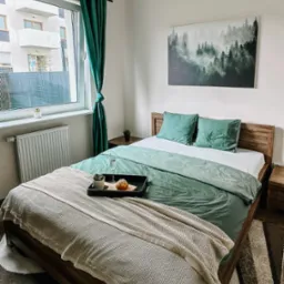 Jasna sypialnia z drewnianym łóżkiem, zieloną pościelą i dekoracją z motywem lasu na ścianie, na łóżku taca ze szklanką wody i bułeczką