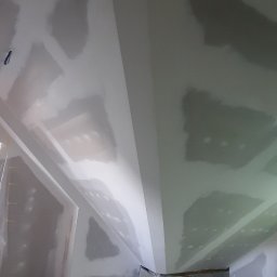 Gauba Ekspert - Poddasze w trakcie prac wykończeniowych: widoczne płyty gipsowo-kartonowe na skośnym suficie, częściowo pokryte szpachlą, z elementem komina obudowanego cegłą u podstawy ściany.