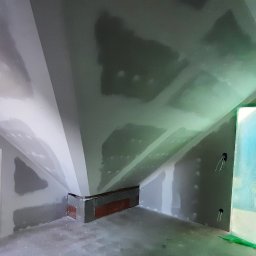 Gauba Ekspert - Poddasze w trakcie prac wykończeniowych: ściany z nałożoną warstwą szpachli, widoczne elementy konstrukcyjne dachu, okno zabezpieczone folią malarską, fragment ściany z cegły i kable elektryczne.