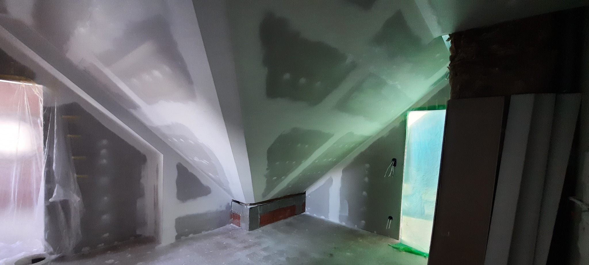 Poddasze w trakcie prac wykończeniowych: ściany z nałożoną warstwą szpachli, widoczne elementy konstrukcyjne dachu, okno zabezpieczone folią malarską, fragment ściany z cegły i kable elektryczne.