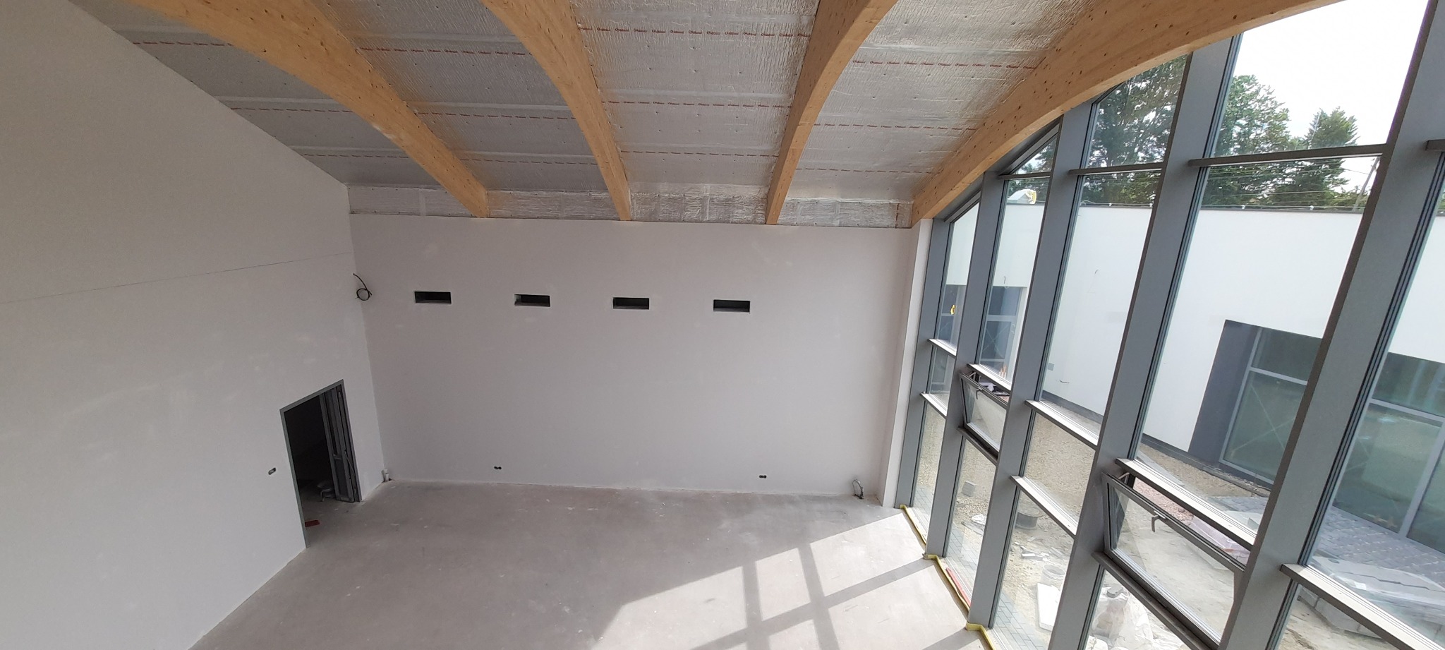 Wnętrze budynku w stanie deweloperskim z drewnianą konstrukcją dachu, ocieplonym stropem, ścianami przygotowanymi pod wykończenie i dużymi oknami.