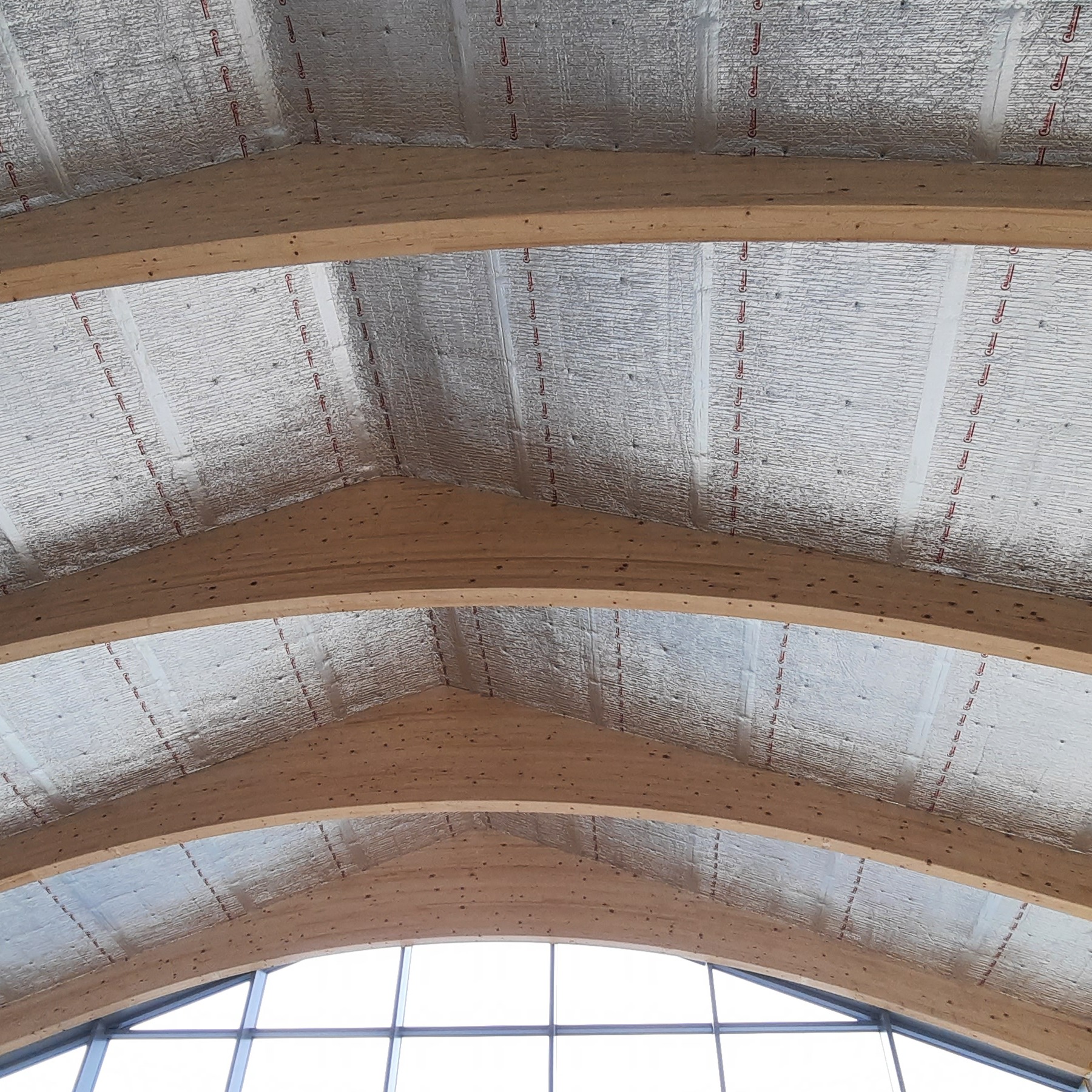 Srebrna folia izolacyjna na drewnianej konstrukcji dachu, widoczna od wewnątrz, z elementami okiennymi w tle.