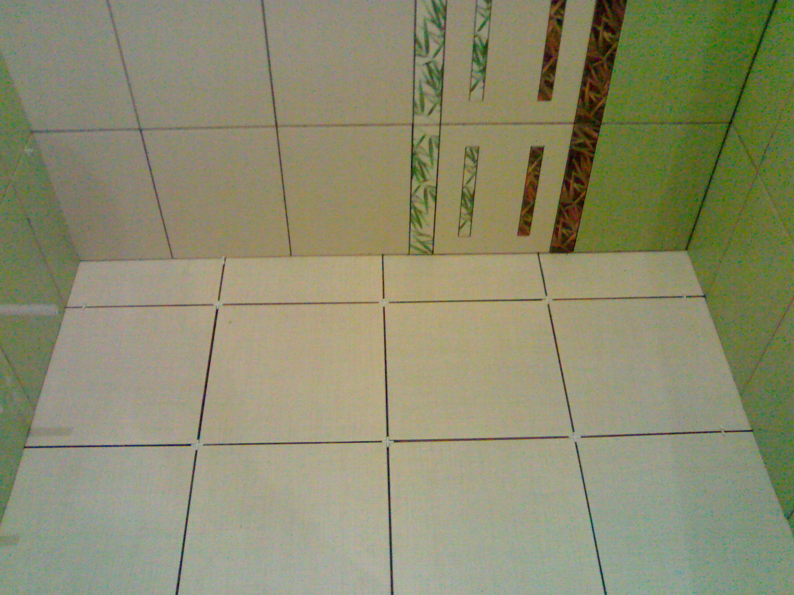 Świeżo ułożone, jasne płytki podłogowe z krzyżykami dystansowymi, widoczne fragmenty dekoracyjnych płytek ściennych z motywami roślinnymi w odcieniach zieleni i brązu.