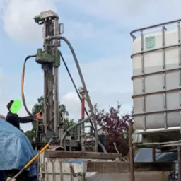 Pracownik obsługujący wiertnicę na zewnątrz, obok zbiorniki na wodę, w tle widoczne drzewa i niebo.