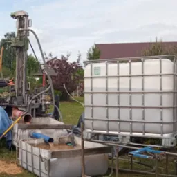 Wiercenie studni: wiertnica w trakcie pracy, woda z urobkiem spływa do zbiorników, widoczny pracownik obsługujący maszynę i duży zbiornik na wodę w tle.