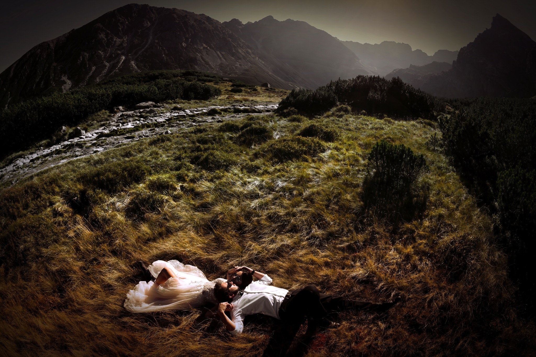 Para młoda leży na trawie na tle gór; panna młoda w białej sukni, pan młody w białej koszuli i czarnych spodniach.