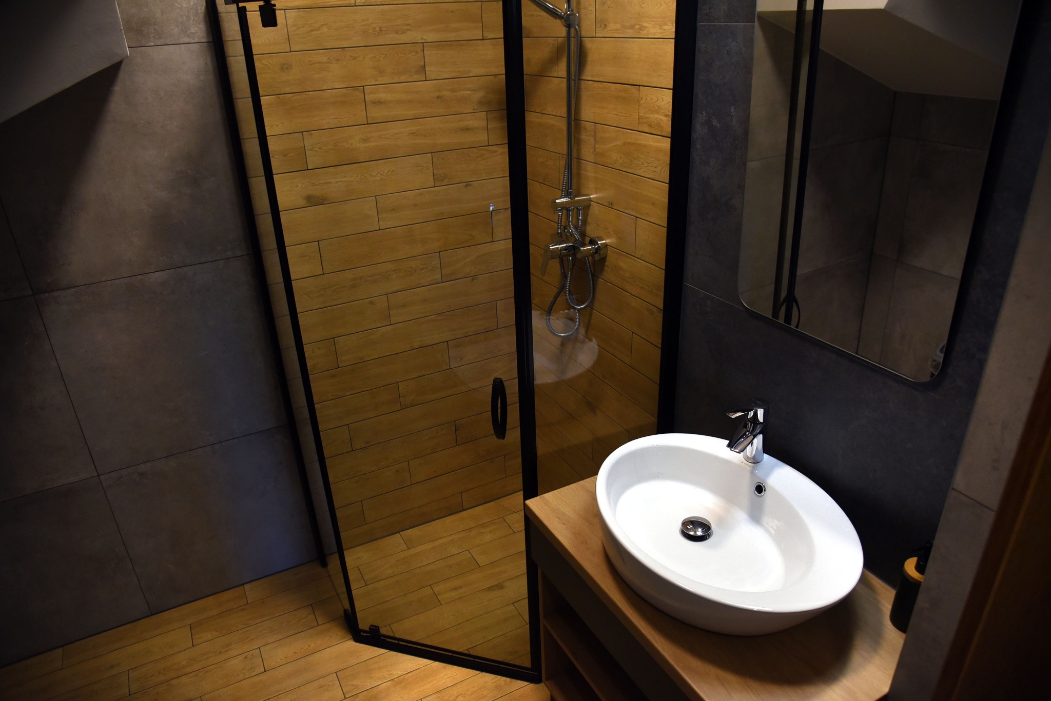 Nowoczesna łazienka z kabiną prysznicową typu walk-in z czarną ramą, drewnopodobnymi płytkami i owalną umywalką nablatową na drewnianym blacie.