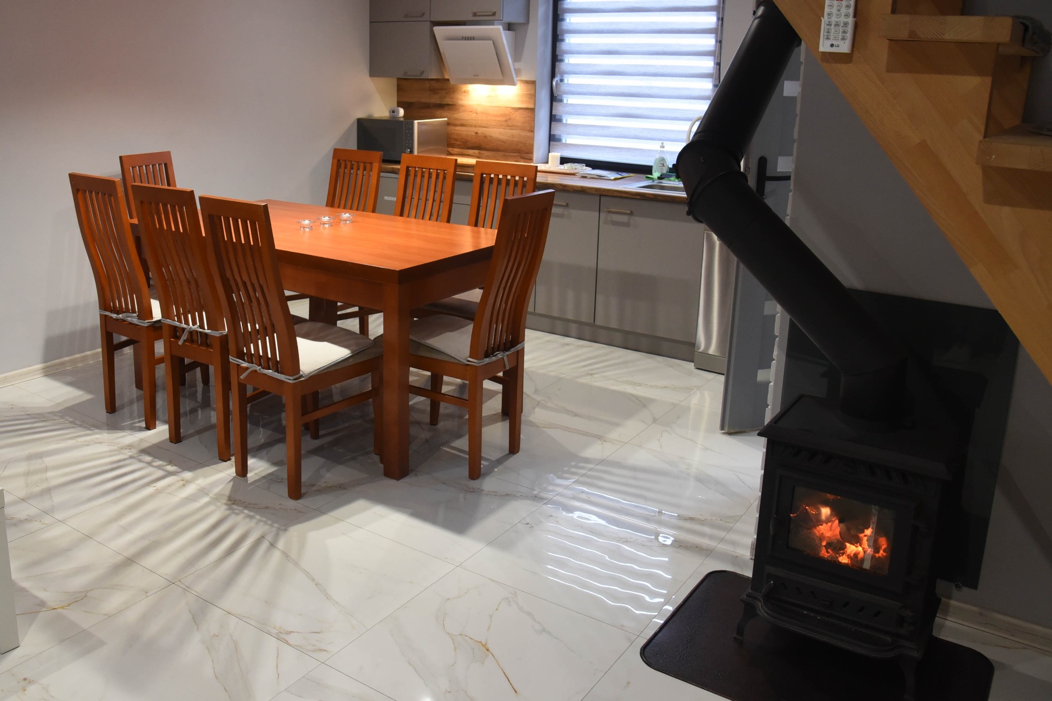 Jasna jadalnia z drewnianym stołem, krzesłami z poduszkami, marmurową podłogą i czarnym piecem kominkowym z widocznym ogniem.