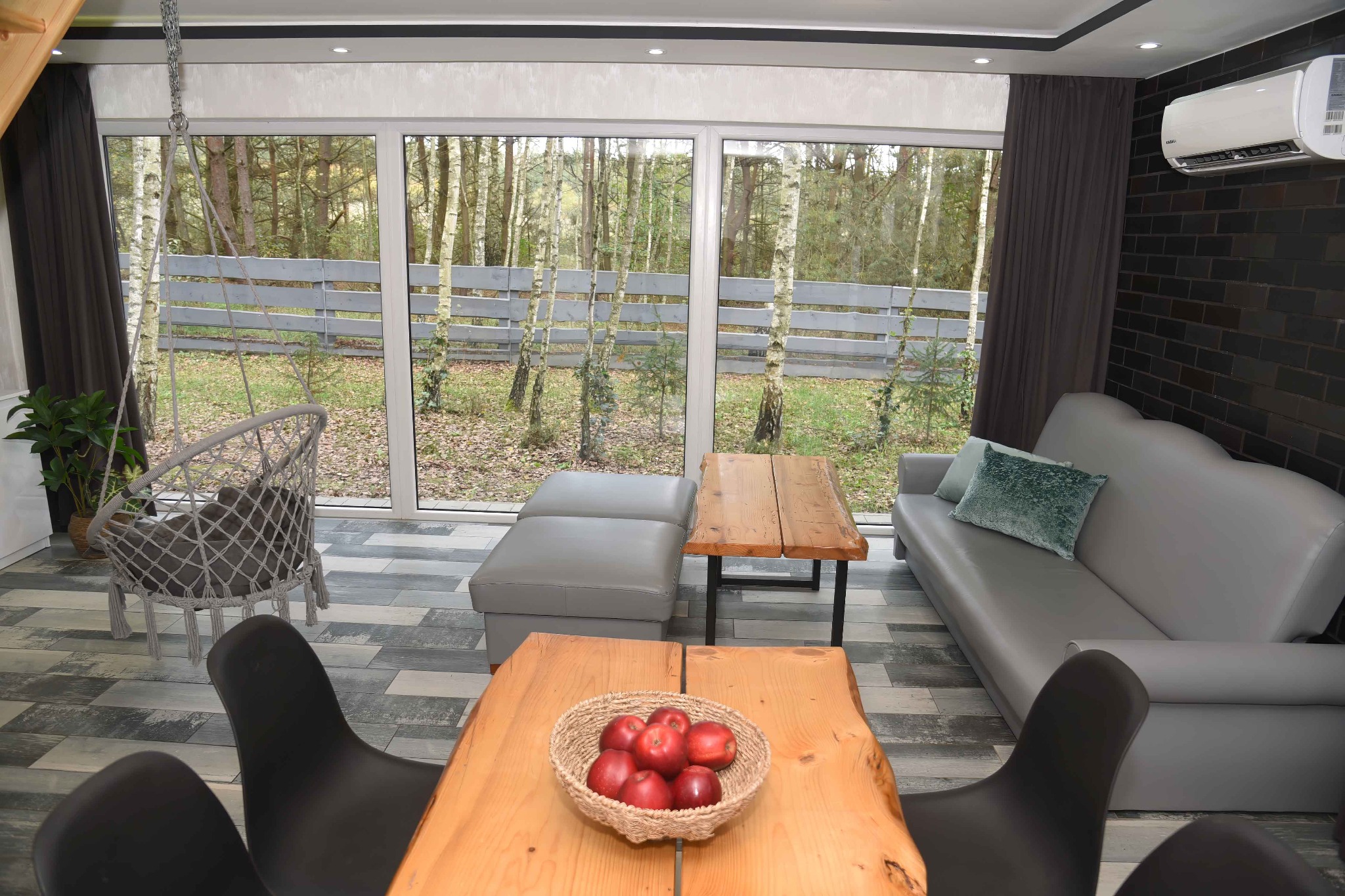 Jasne wnętrze salonu z dużym oknem na las, drewnianymi stołami, szarą sofą i wiszącym fotelem, kosz z czerwonymi jabłkami na pierwszym planie.