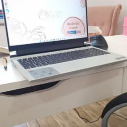 Laptop Dell na białym biurku z widocznym logo Bloom Media na ekranie, obok leży ergonomiczna myszka i długopis, w tle krzesło i fragment pokoju.
