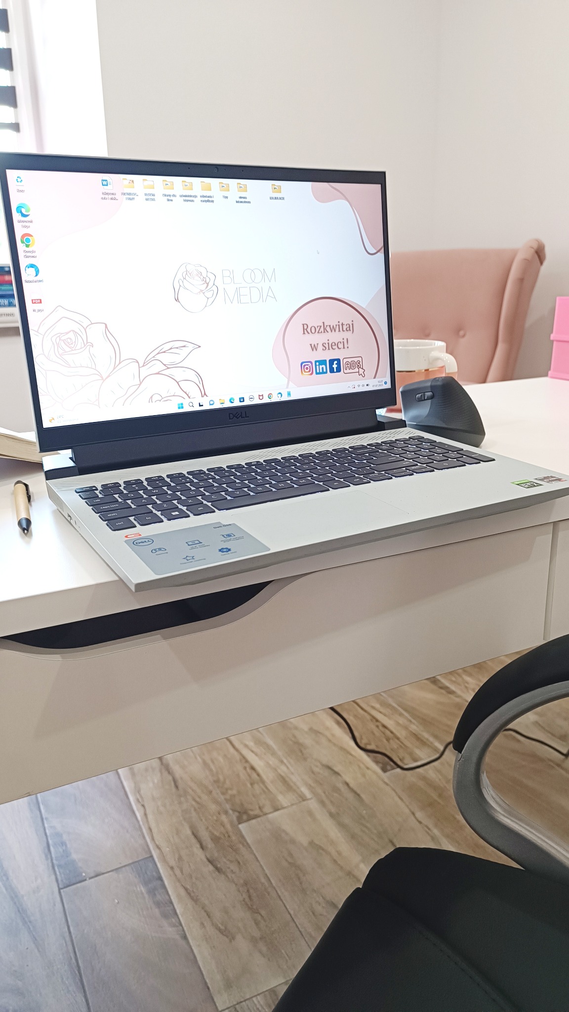 Laptop Dell na białym biurku z widocznym logo Bloom Media na ekranie, obok leży ergonomiczna myszka i długopis, w tle krzesło i fragment pokoju.