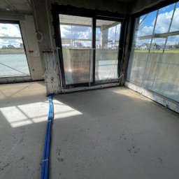 El-Dom Dominik Szulc - Wnętrze surowego budynku w trakcie wykańczania, widoczne niebieskie rury instalacyjne prowadzone wzdłuż ściany, wystające przewody elektryczne oraz duże okna z widokiem na zabudowę jednorodzinną.