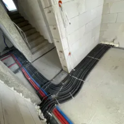 Rury instalacyjne w peszlach ułożone równolegle na surowym podłożu w nowo budowanym budynku, zabezpieczone metalowymi klamrami, widoczne przewody elektryczne w ścianie z betonu komórkowego.