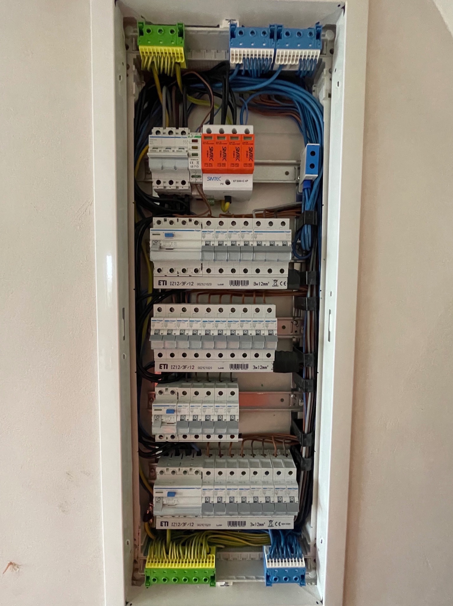 Otwarta skrzynka elektryczna z bezpiecznikami, przewodami w różnych kolorach i listwami zaciskowymi, widoczne elementy SIMTEC i ETI, zamontowana na jasnej ścianie.