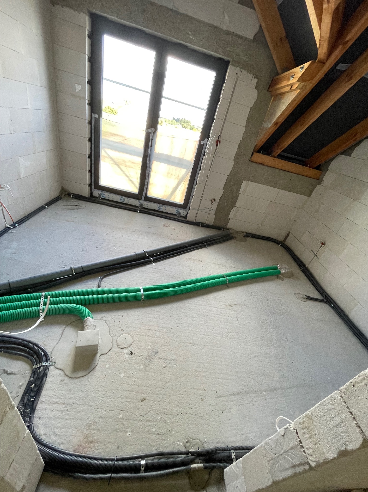 Wnętrze w stanie surowym z widocznymi przewodami elektrycznymi i rurami w peszlach, zamocowanymi do ściany i podłogi, okno dachowe w tle.