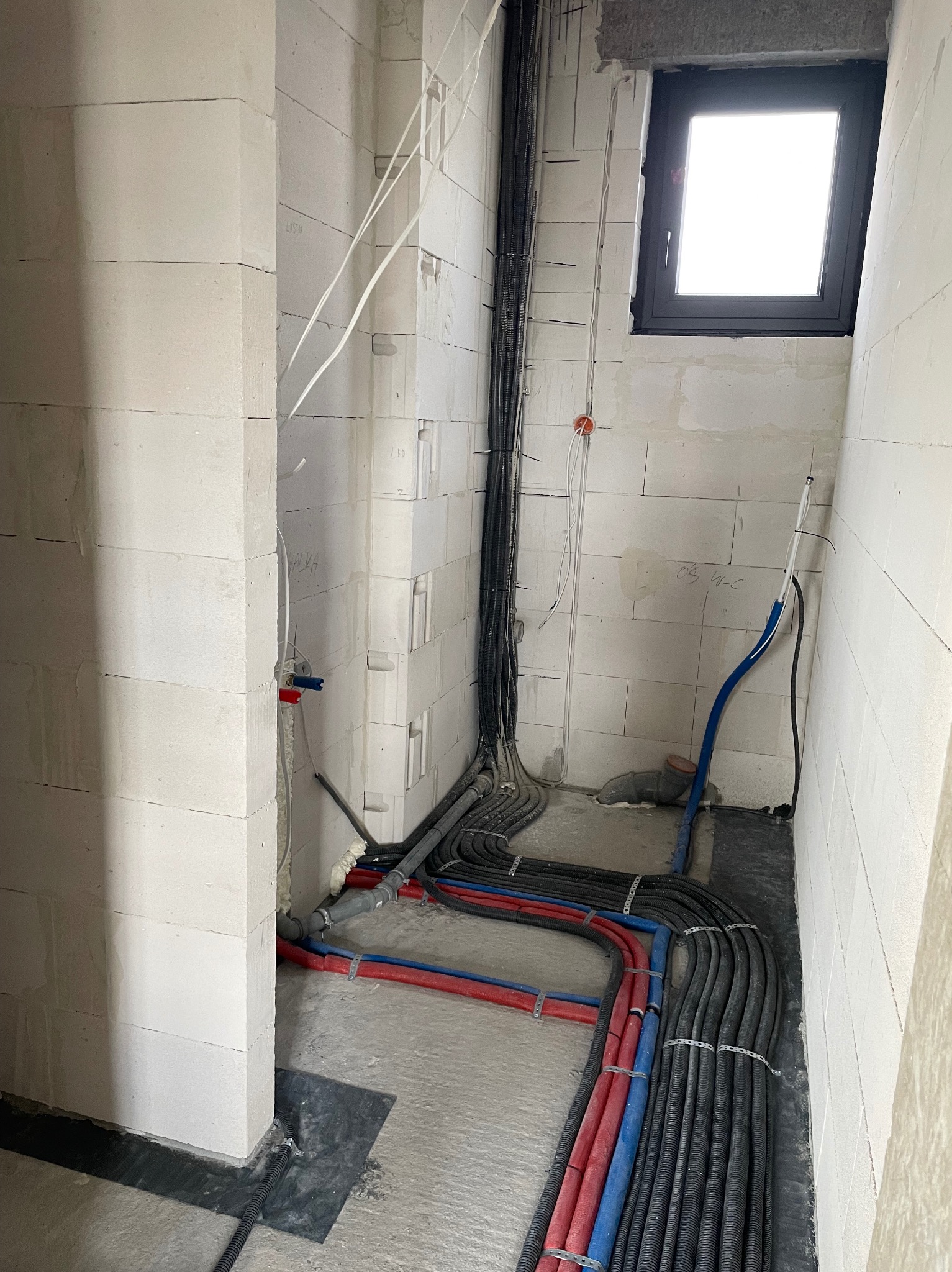 Surowy stan instalacji elektrycznej i hydraulicznej w nowym pomieszczeniu z białych bloczków, widoczne rury grzewcze czerwone i niebieskie, czarne kable w peszlach, okno w czarnej ramie...