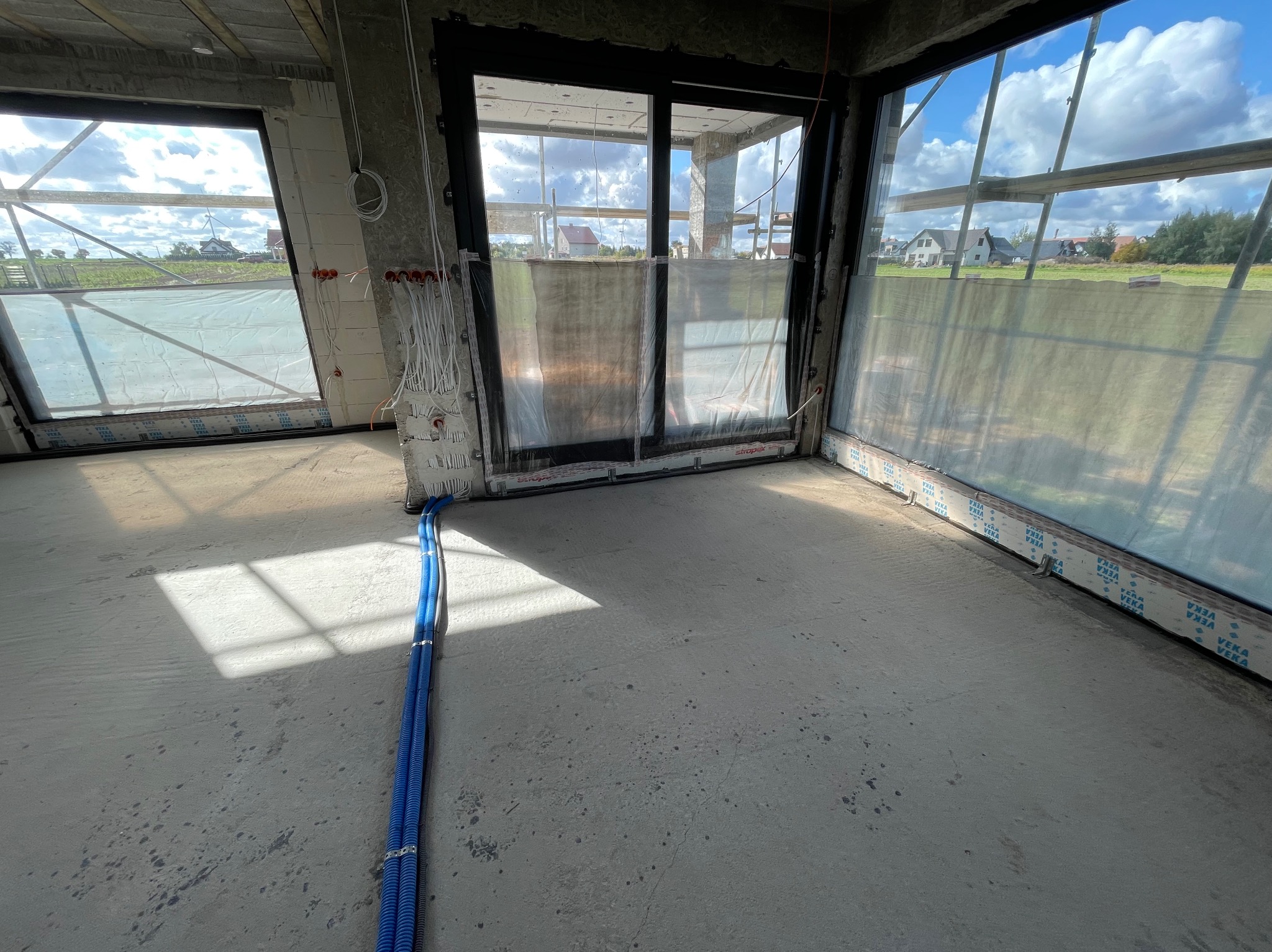 Wnętrze surowego budynku w trakcie wykańczania, widoczne niebieskie rury instalacyjne prowadzone wzdłuż ściany, wystające przewody elektryczne oraz duże okna z widokiem na zabudowę jednorodzinną.