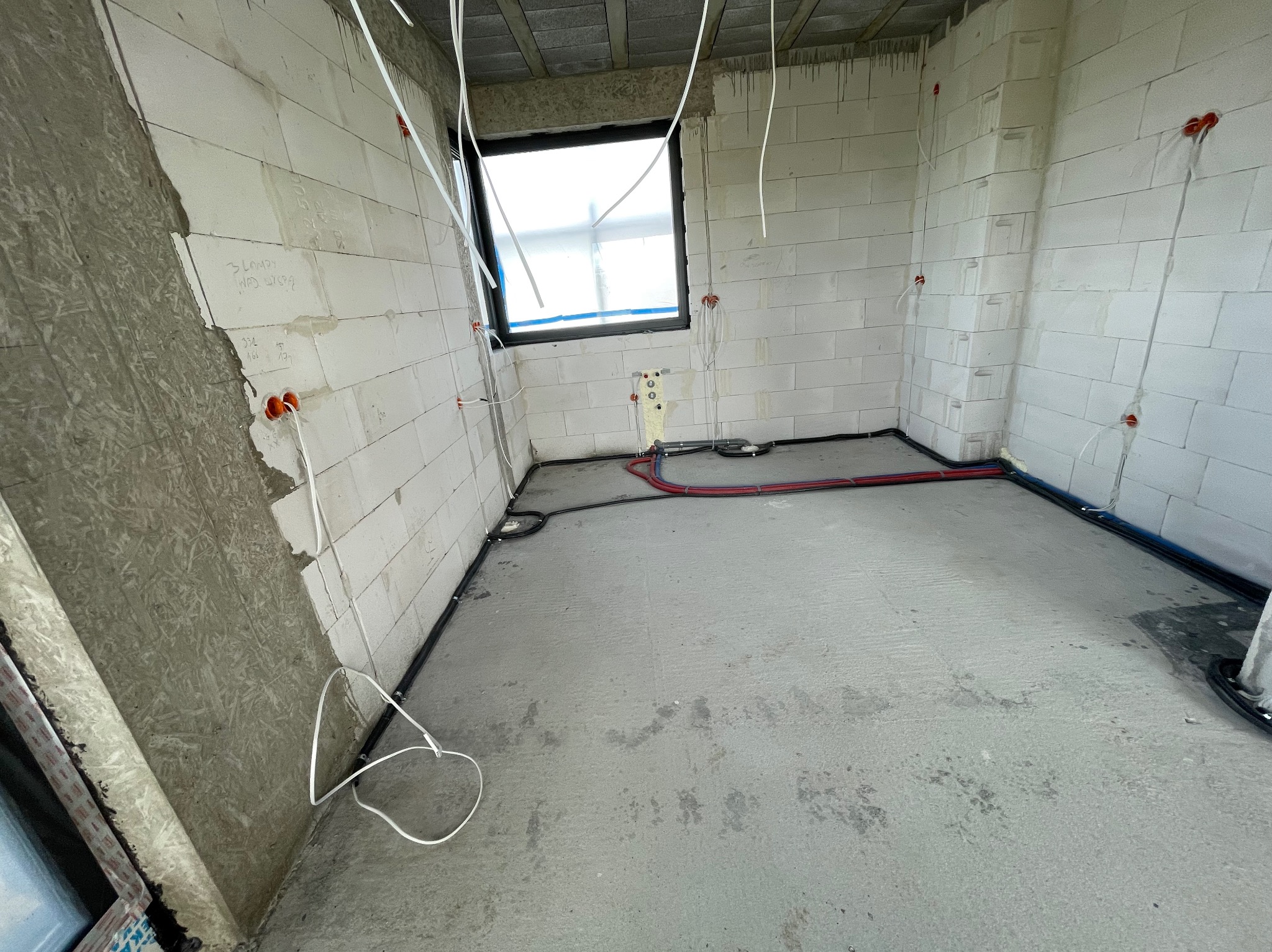 Surowy stan wnętrza z widoczną instalacją elektryczną w ścianach i podłodze, przewody wystające z puszek, ściany z betonu komórkowego, okno zabezpieczone folią.