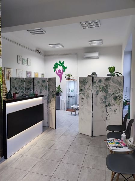 Wnętrze gabinetu z recepcją, parawanem z motywem liści i logo z sylwetką człowieka otoczoną zielonymi liśćmi, na ścianie wiszą ramki z dyplomami.