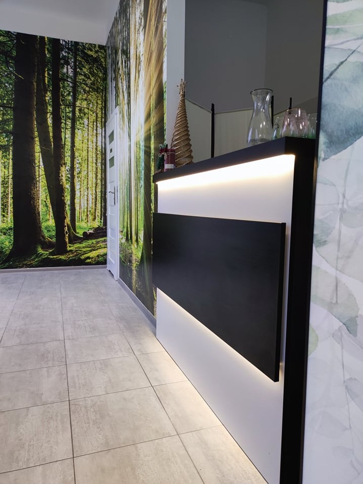 Nowoczesna recepcja z podświetleniem LED, dekoracyjnym drzewkiem i fototapetą leśną w tle, w jasnym wnętrzu.