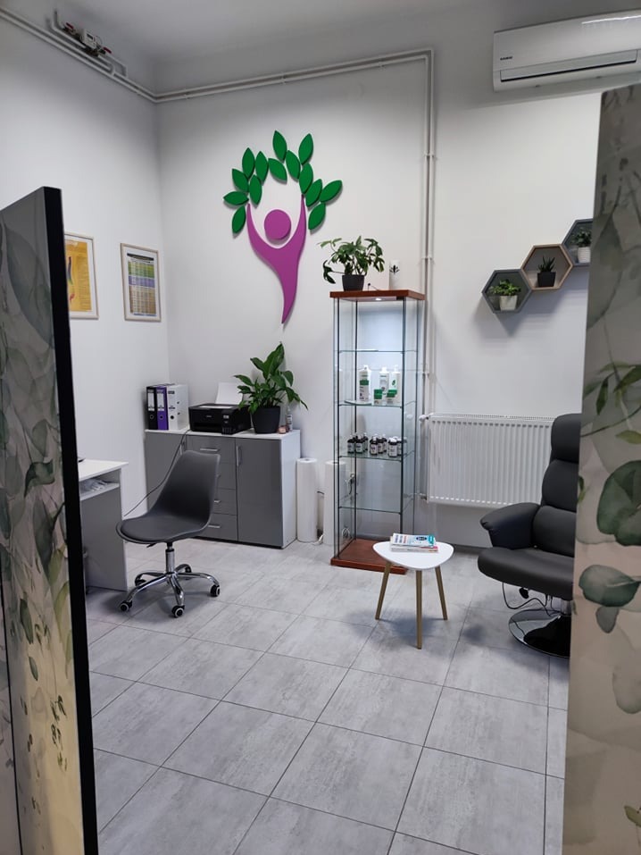 Jasne wnętrze gabinetu z fotelem, biurkiem, szafką z dokumentami, szklaną witryną z produktami i dekoracją ścienną w kształcie osoby otoczonej liśćmi, sugerujące holistyczne podejście do zdrowia.