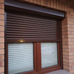 Ciemnobrązowa osłona okienna zewnętrzna, częściowo opuszczona, na tle ściany z cegły. W oknach widoczne białe plisy.