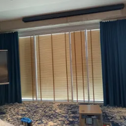 Beżowe poziome lamele z drewnianymi akcentami w oknach, zasłonięte niebieskimi zasłonami w jasnym pomieszczeniu z wzorzystą wykładziną.