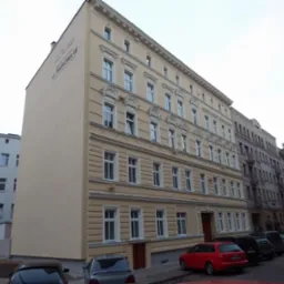 Odnowiona elewacja kamienicy w beżowych barwach, z widocznymi detalami architektonicznymi i zaparkowanymi autami. Budynek po remoncie, widok z ulicy.