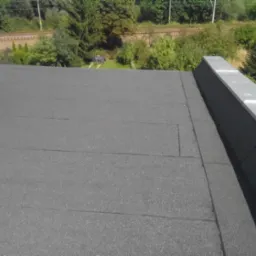 Wykończony, płaski dach pokryty papą termozgrzewalną. Widoczne łączenia papy i obróbki blacharskie. W tle zieleń i fragment infrastruktury kolejowej.