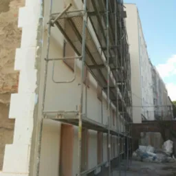 Ocieplanie budynku z użyciem rusztowania. Widoczny fragment elewacji z ociepleniem w postaci białych płyt oraz fragment ściany przed ociepleniem. Prace w toku.